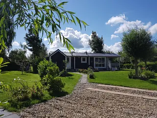 Een klein huis met één verdieping, een blauw dak, witte muren met rode accenten en een voorterras met stoelen, omringd door groen