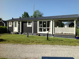 Een klein huis met één verdieping, een blauw dak, witte muren met rode accenten en een voorterras met stoelen, omringd door groen